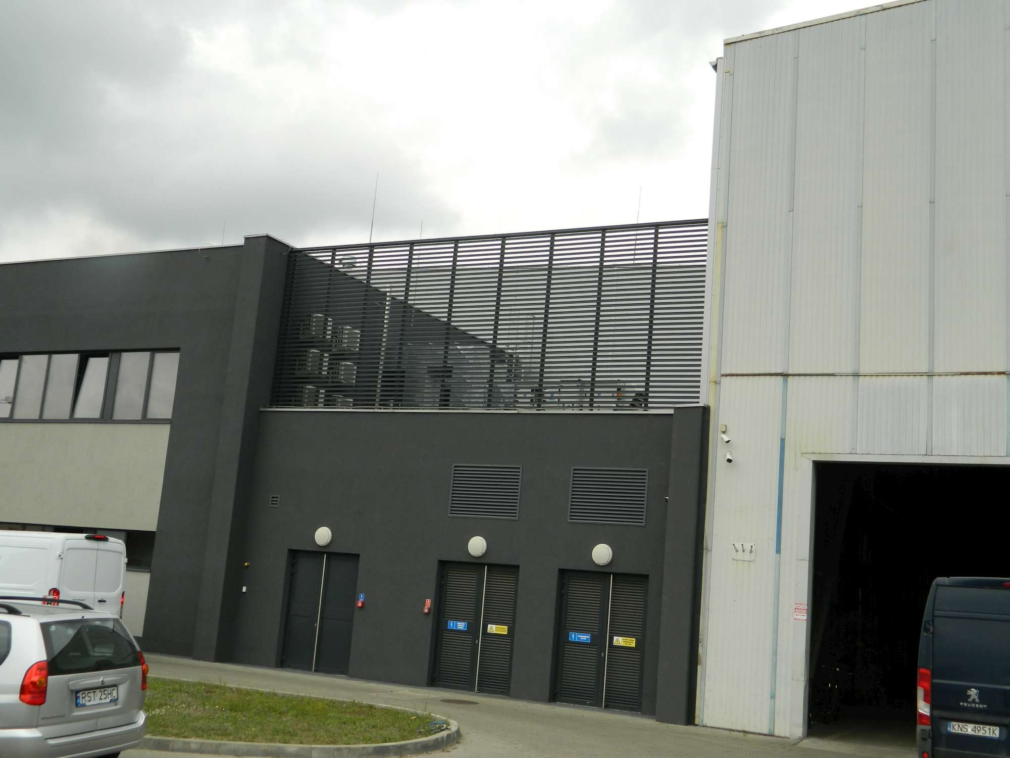 Szary budynek przemysłowy z trzema parami drzwi wentylacyjnych, widoczne samochody dostawcze i osobowe na parkingu, pochmurne niebo.