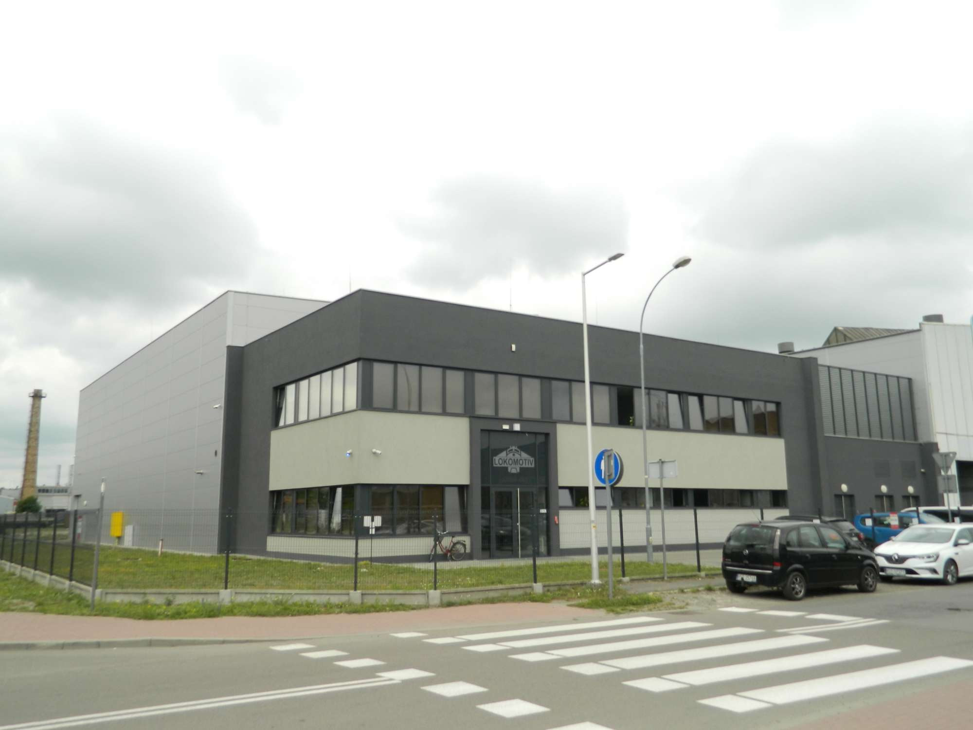 Nowoczesny budynek biurowy z szaro-beżową elewacją, widok z ulicy z przejściem dla pieszych i zaparkowanymi samochodami, w tle komin przemysłowy.