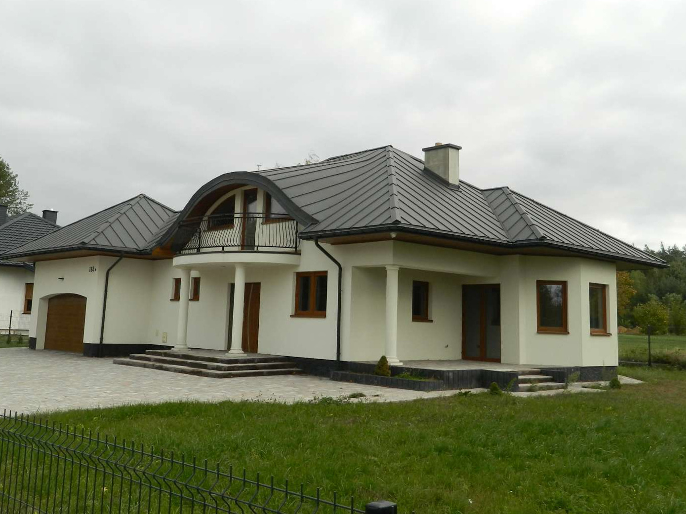 Nowoczesny, parterowy dom z grafitowym dachem blaszanym, białymi ścianami i kolumnami podtrzymującymi balkon, widoczny zza czarnego, metalowego ogrodzenia na tle pochmurnego nieba.
