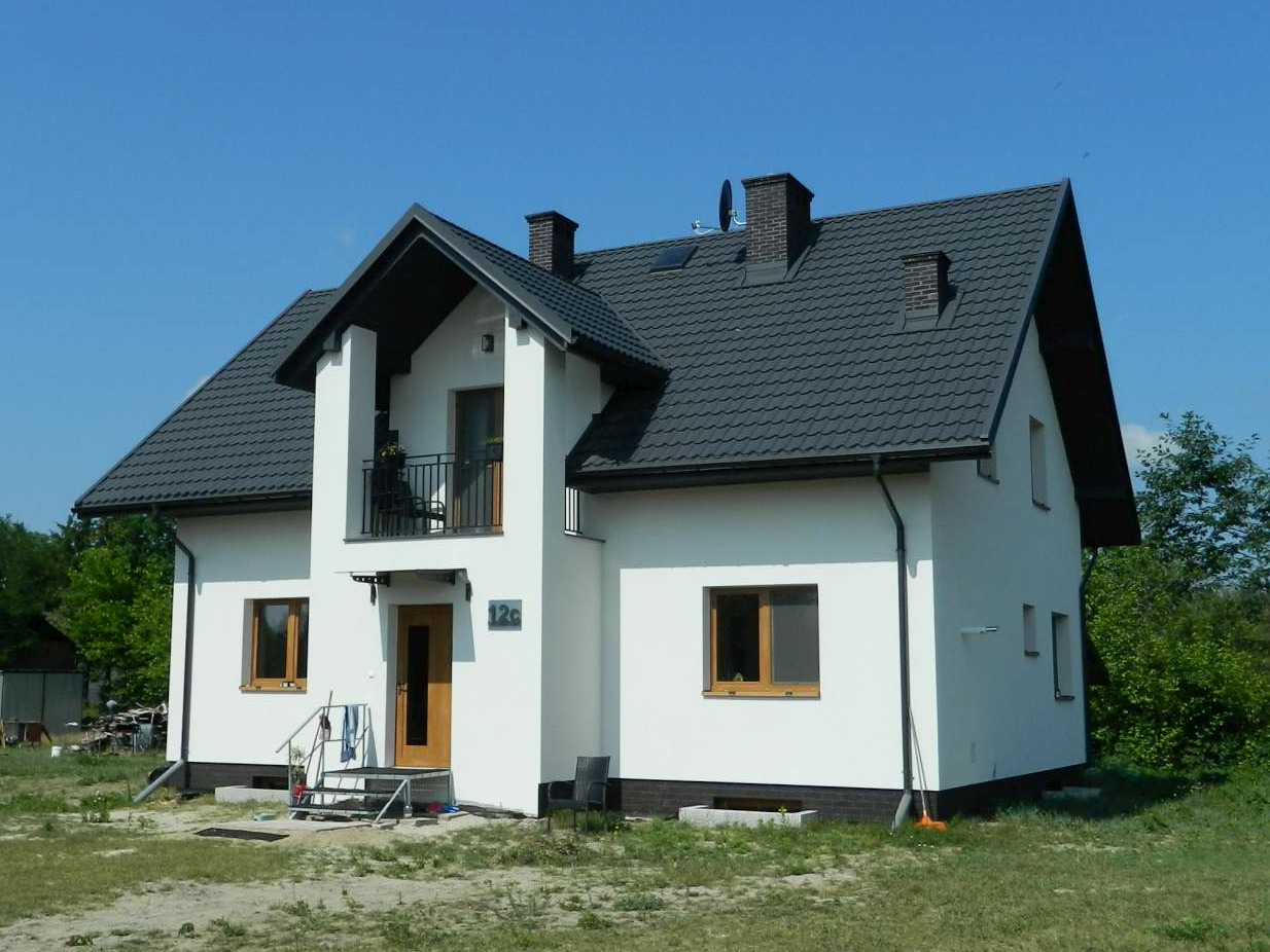 Nowoczesny, dwukondygnacyjny dom jednorodzinny z szarą dachówką i białymi ścianami, numer 12c nad drzwiami, widok z zewnątrz w słoneczny dzień.