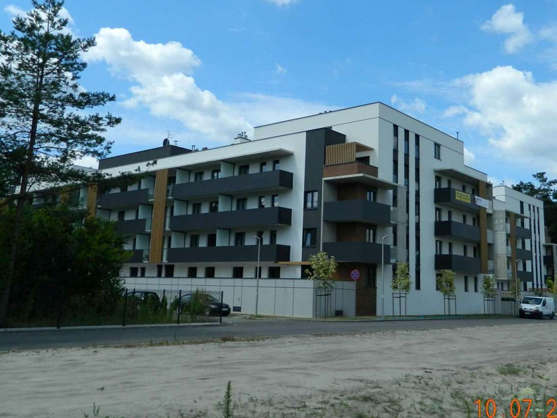 Nowoczesny, wielopiętrowy budynek mieszkalny z balkonami w odcieniach szarości i bieli, widoczny z perspektywy ulicy, z drzewem po lewej stronie i samochodami w tle.