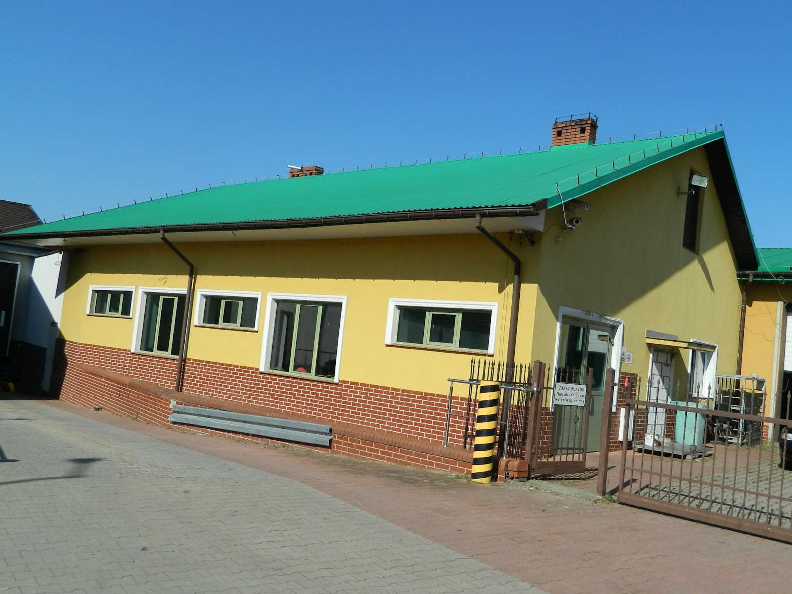 Żółty budynek z zielonym dachem, fragment muru z cegły, widok z zewnątrz, brama wjazdowa.