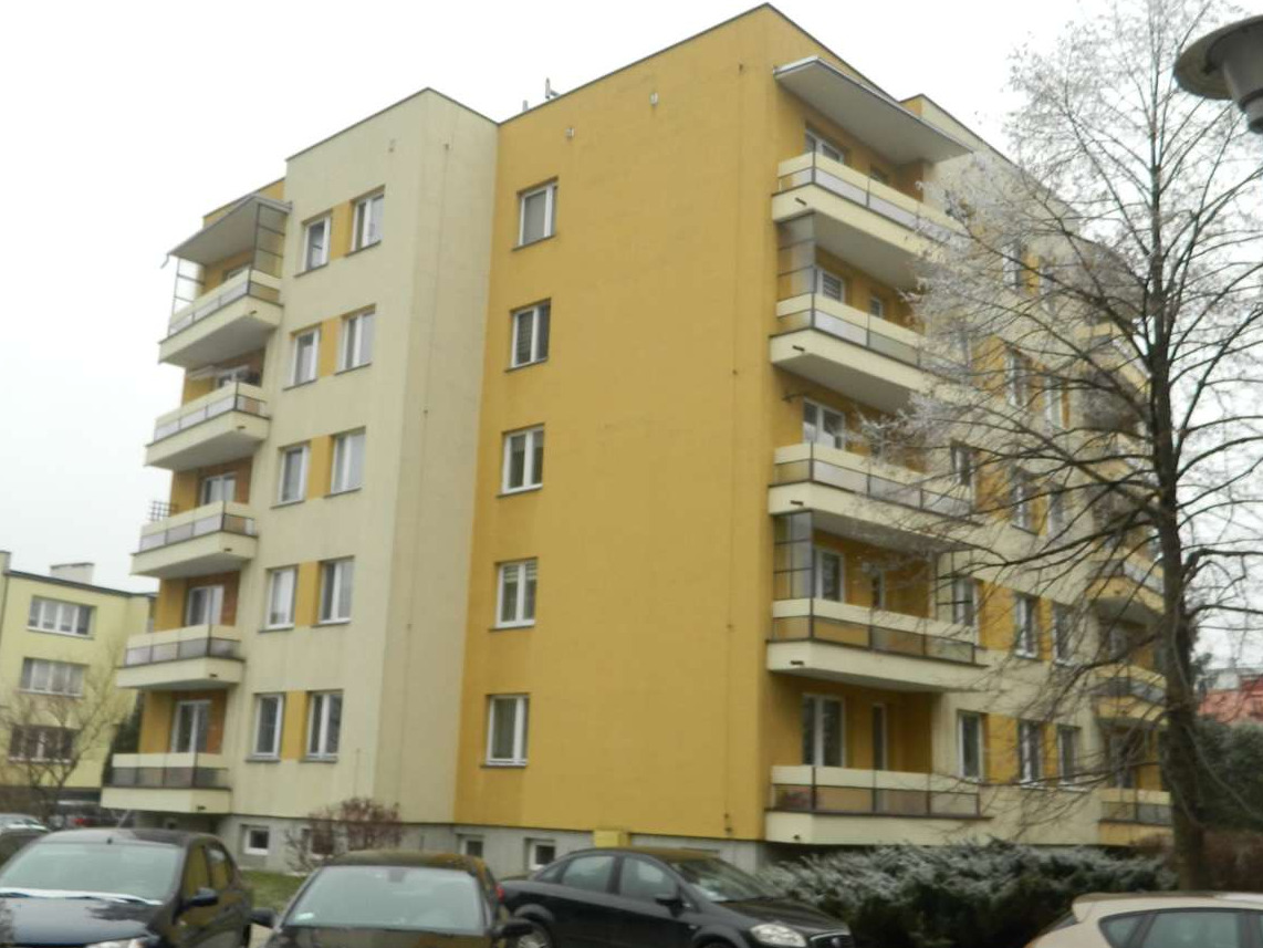 Żółto-beżowy blok mieszkalny z balkonami, widok z dołu, pochmurne niebo, samochody na parkingu