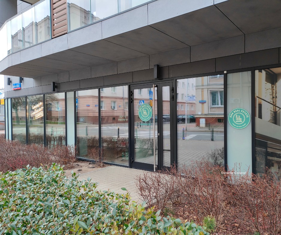 Fasada biura agencji nieruchomości z logo 'AHA Real Estate' na szybie i neonem w oknie, otoczona zielenią i chodnikiem.