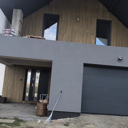GUM-BUD - Nowoczesny dom jednorodzinny w Podedwórzu z szarą elewacją, drewnianą okładziną i grafitową bramą garażową. Widoczne worki z materiałami budowlanymi i miotła.