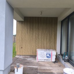 GUM-BUD - Wykończona elewacja z drewnianą okładziną na zadaszonym tarasie. Widoczny styropian, narzędzia i wiadro z farbą. Częściowo widoczne okno i kolumna.