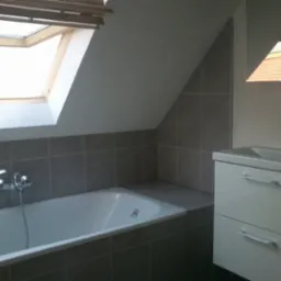 Szara łazienka na poddaszu z oknem dachowym, biała wanna i umywalka z szafką, płytki ceramiczne na ścianach.