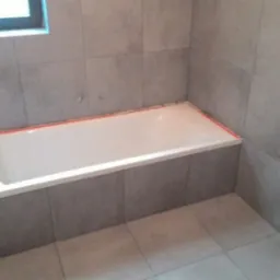 Biała wanna obudowana szarymi płytkami w trakcie montażu, widoczne zabezpieczenia krawędzi pomarańczową taśmą malarską, szare płytki na ścianach i podłodze, okno w górnym lewym rogu kadru.