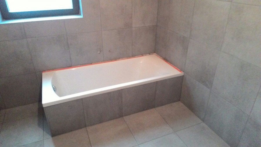 Biała wanna obudowana szarymi płytkami w trakcie montażu, widoczne zabezpieczenia krawędzi pomarańczową taśmą malarską, szare płytki na ścianach i podłodze, okno w górnym lewym rogu kadru.