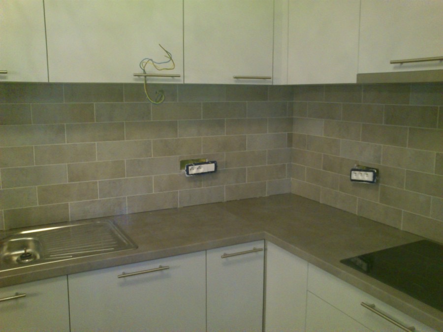 Kuchnia w trakcie wykańczania: ułożone szare płytki imitujące cegłę na ścianie nad blatem, widoczne gniazdka elektryczne przed montażem osprzętu oraz wystające przewody elektryczne.