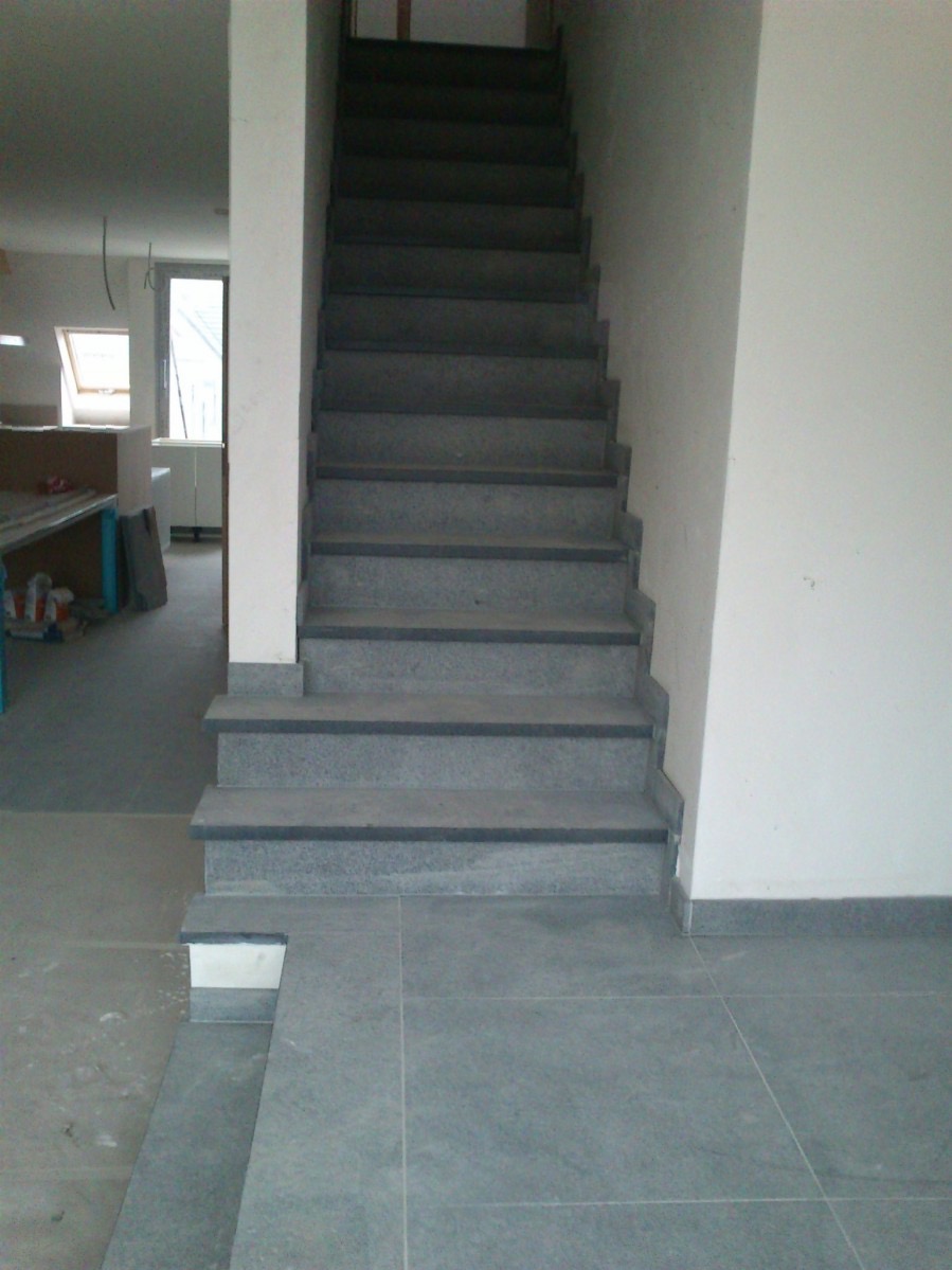 Wykończone schody z szarego kamienia prowadzące na górę, z szarą podłogą z płytek i widocznym fragmentem pomieszczenia w tle.
