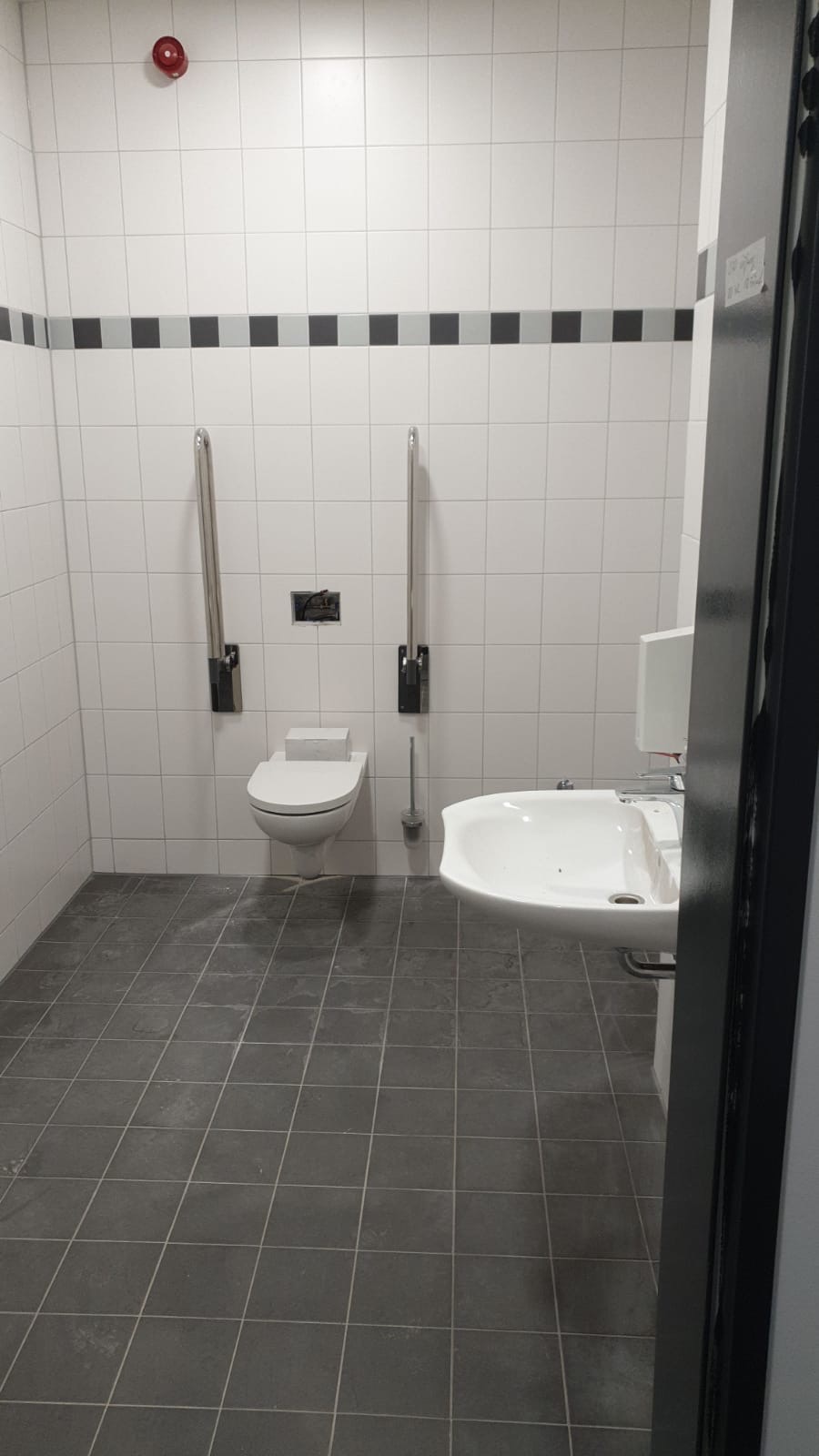 Wykończona łazienka dla osób z niepełnosprawnościami: białe płytki ścienne z dekoracyjnym pasem, szare płytki podłogowe, toaleta z uchwytami, umywalka. Widoczny system alarmowy na suficie.