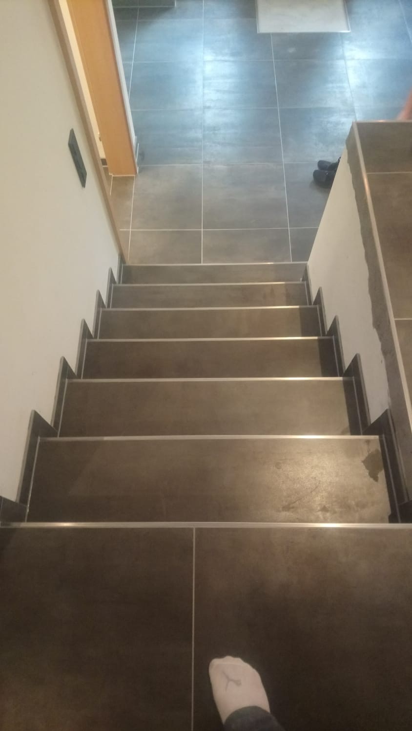Widok z góry na wykończone schody z ciemnych płytek z metalowymi listwami na krawędziach, prowadzące do pomieszczenia z szarą podłogą wyłożoną płytkami. Na dole kadru widoczna stopa w skarpetce.