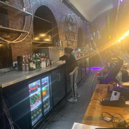 EcoSpark.Pro - Pracownik w czarnej bluzie i szarych spodniach wyciera bar w lokalu z ceglanymi ścianami, widoczne butelki alkoholi, chłodziarka z napojami, terminal płatniczy i osoba na drabinie w tle.