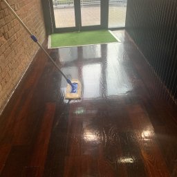 EcoSpark.Pro - Mop z niebieskimi akcentami czyści błyszczącą, ciemną drewnianą podłogę w pobliżu drzwi wejściowych z zieloną wycieraczką i ceglanej ściany.