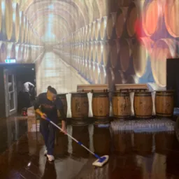 Mężczyzna z mopem czyści błyszczącą, drewnianą podłogę w pomieszczeniu z dekoracją ścienną imitującą piwnicę z beczkami, widoczne pudełka i szklanki w tle.