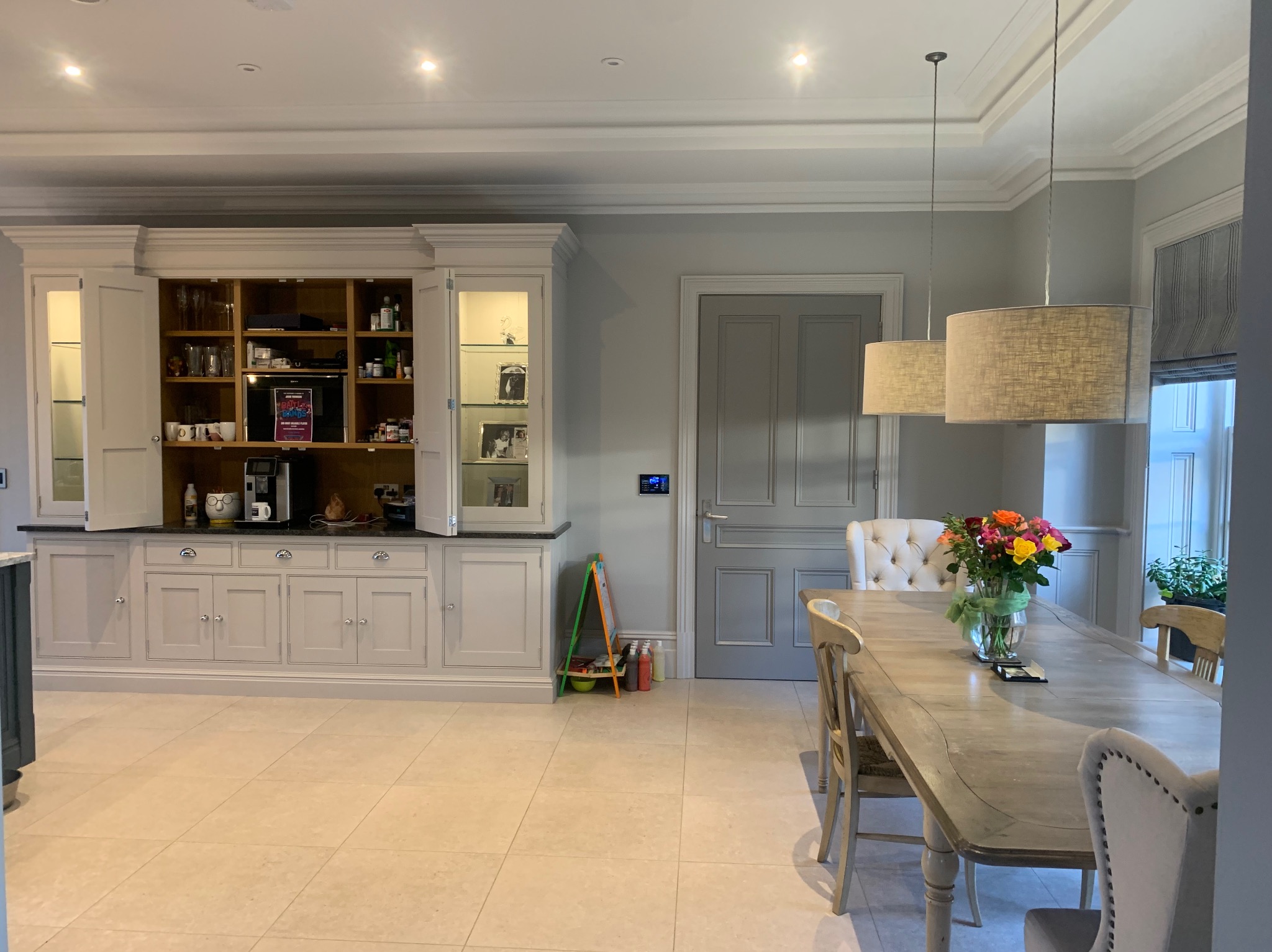 Jasne wnętrze jadalni z dużym kredensem, drewnianym stołem z bukietem kolorowych kwiatów w wazonie i dwiema lampami wiszącymi z abażurami.