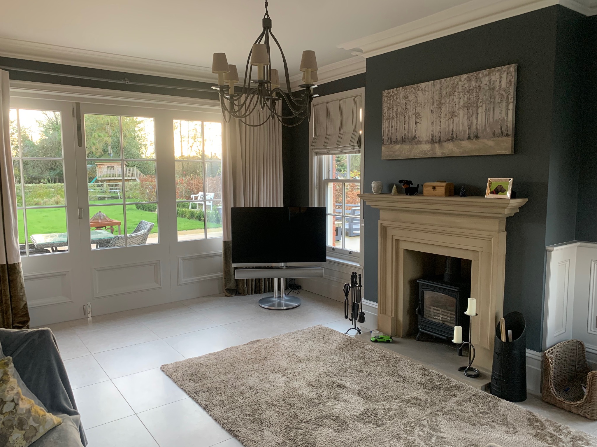 Jasny salon z dużym oknem wychodzącym na ogród, kremową podłogą, beżowym dywanem, kominkiem z czarnym piecem i telewizorem na srebrnej podstawie.