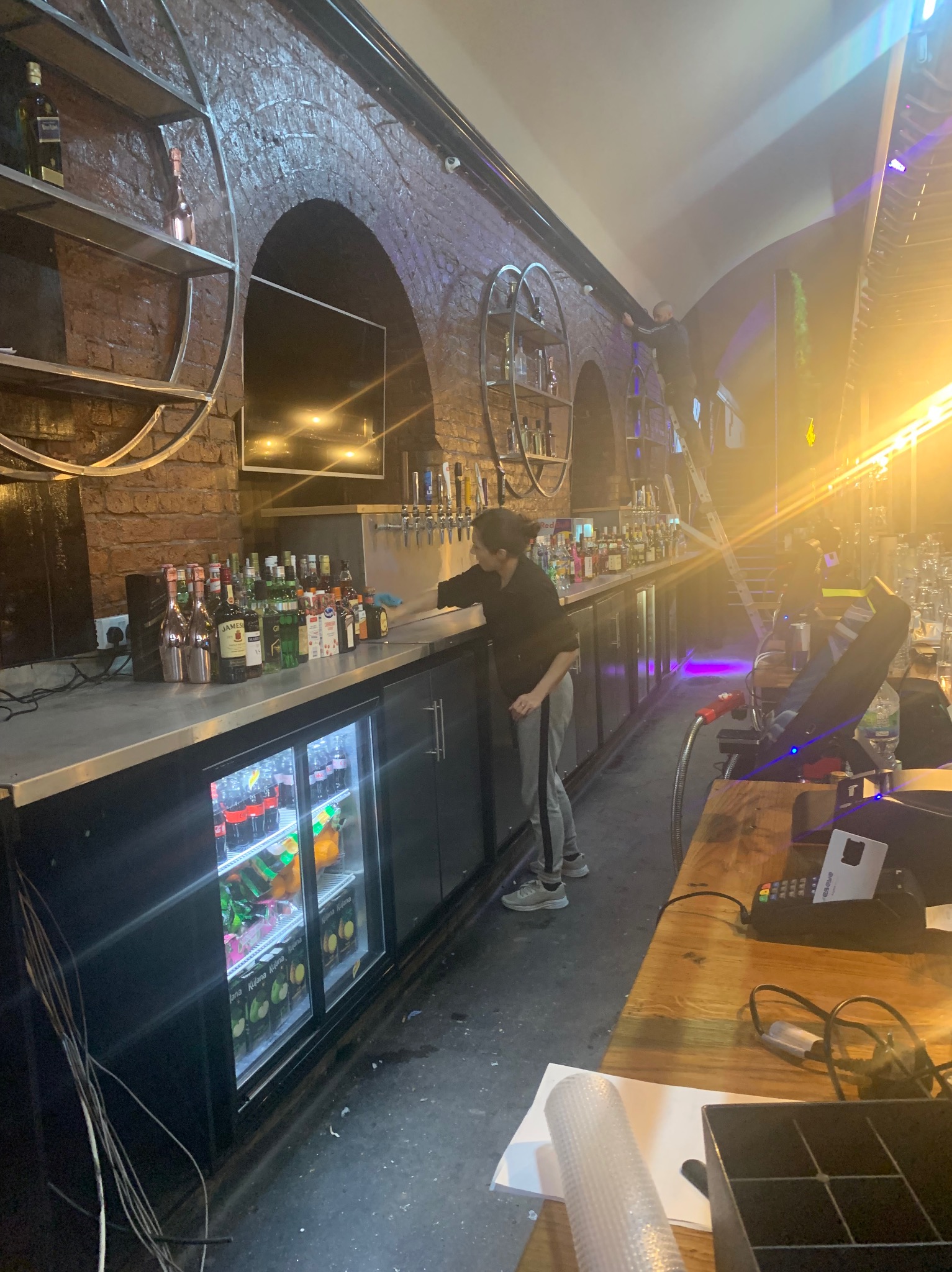 Pracownik w czarnej bluzie i szarych spodniach wyciera bar w lokalu z ceglanymi ścianami, widoczne butelki alkoholi, chłodziarka z napojami, terminal płatniczy i osoba na drabinie w tle.
