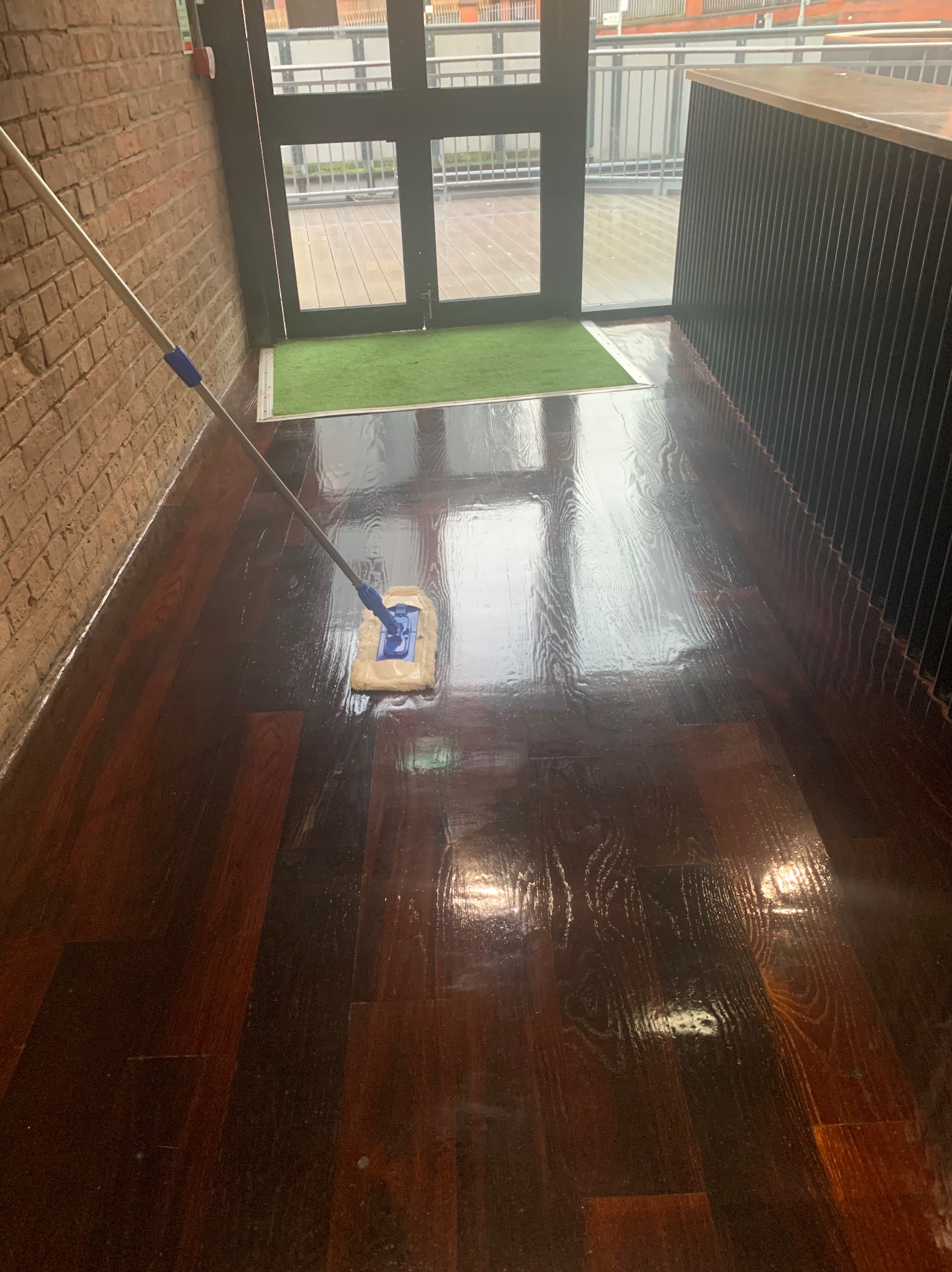 Mop z niebieskimi akcentami czyści błyszczącą, ciemną drewnianą podłogę w pobliżu drzwi wejściowych z zieloną wycieraczką i ceglanej ściany.