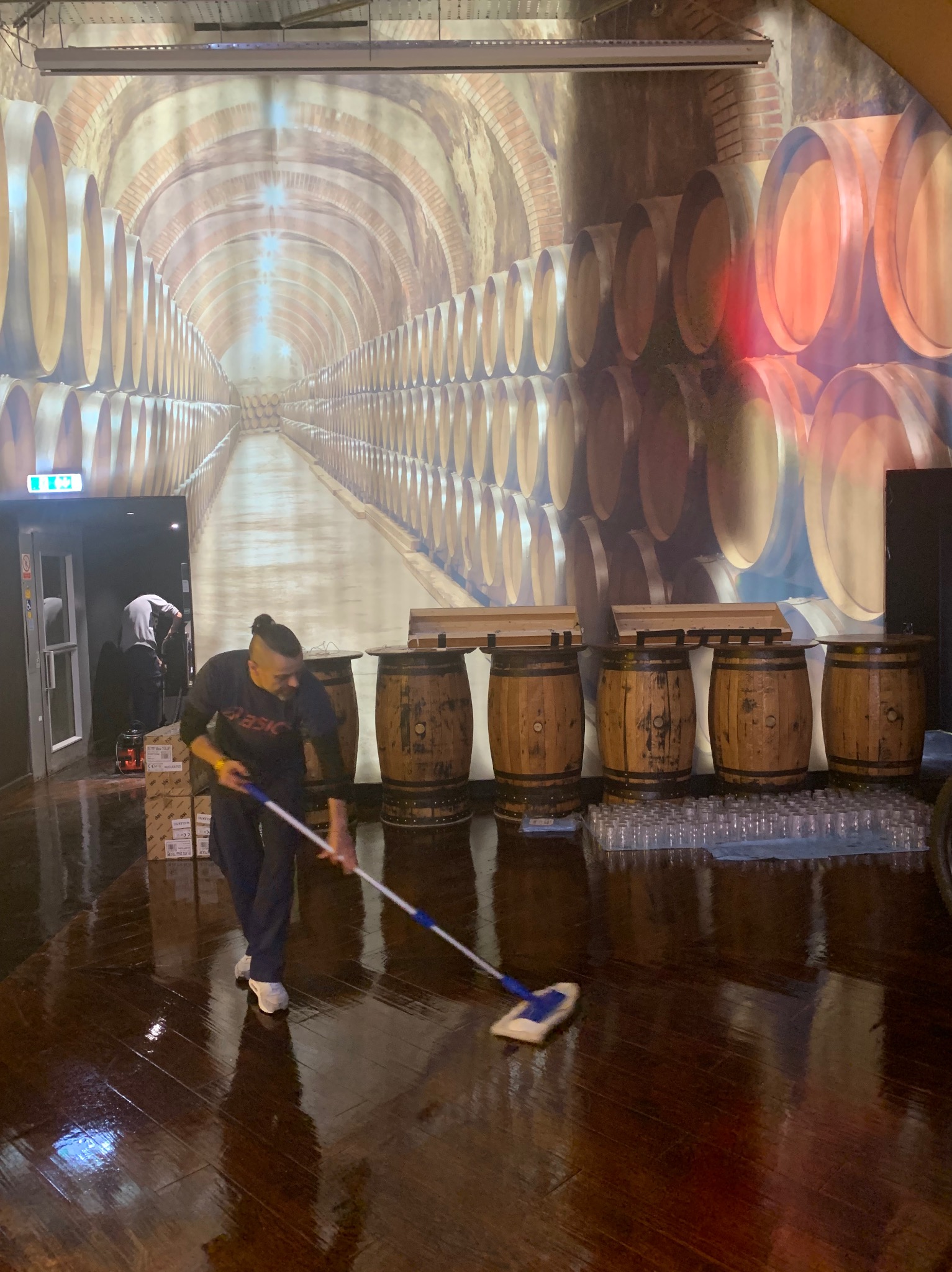 Mężczyzna z mopem czyści błyszczącą, drewnianą podłogę w pomieszczeniu z dekoracją ścienną imitującą piwnicę z beczkami, widoczne pudełka i szklanki w tle.