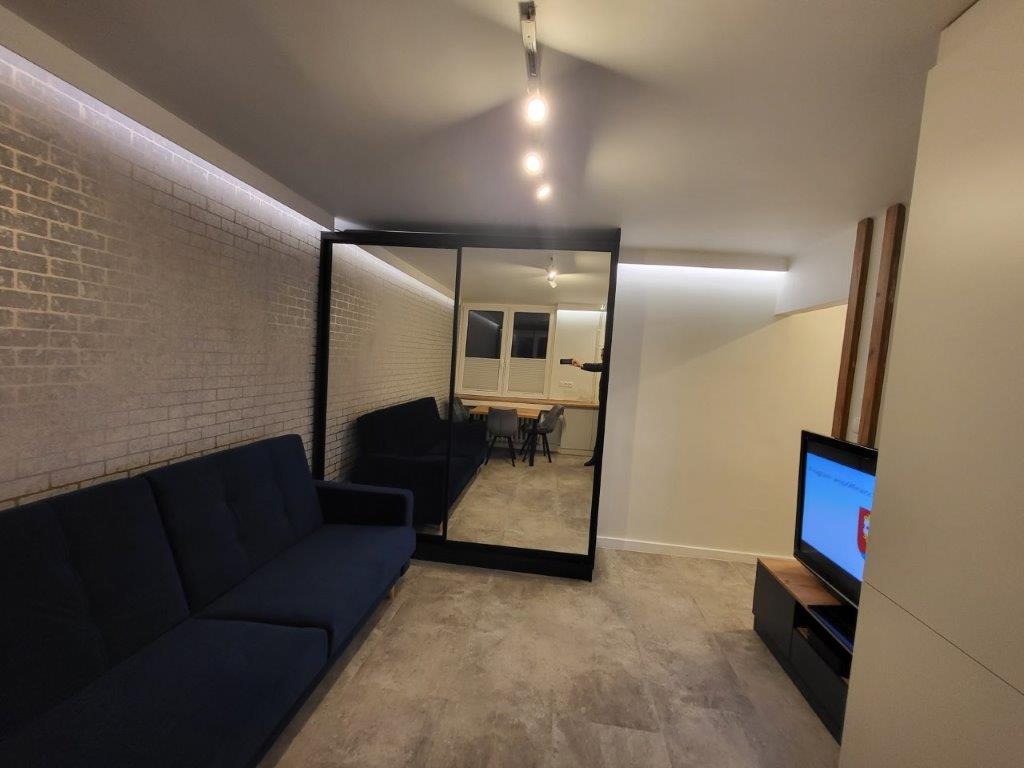 Salon z ciemnoniebieską sofą, ściana z cegieł pomalowana na szaro, duże lustro w czarnej ramie odbijające sofę i stół z krzesłami, telewizor na szafce RTV.