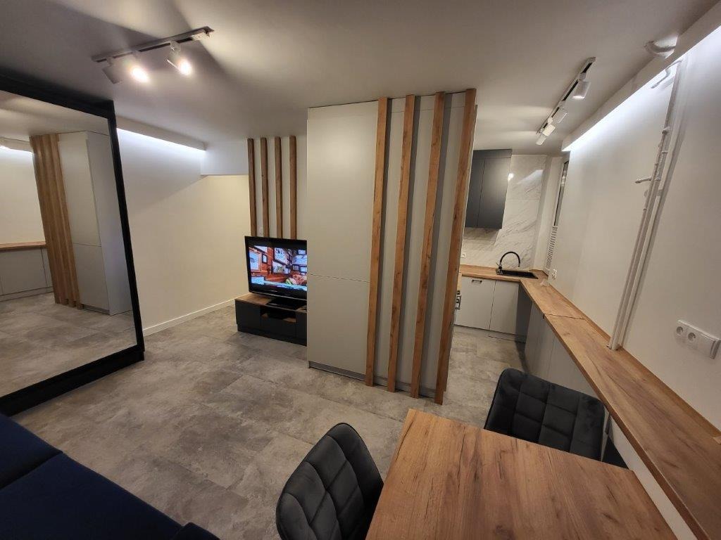 Nowoczesne wnętrze mieszkania z szarymi ścianami, drewnianymi akcentami, kuchnią w tle i dużym lustrem po lewej stronie, widok na stół z krzesłami i telewizor.