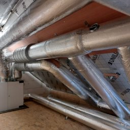 Firma Handlowo-Usługowa Krzysztof Ścibior - System kanałów wentylacyjnych z centralą rekuperacyjną w nowym budynku, srebrna izolacja na rurach, widoczne elementy montażowe.