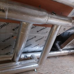 Firma Handlowo-Usługowa Krzysztof Ścibior - Izolowane rury wentylacyjne w przestrzeni poddasza, widoczne elementy konstrukcyjne dachu i izolacja z nadrukiem logo Unilin.