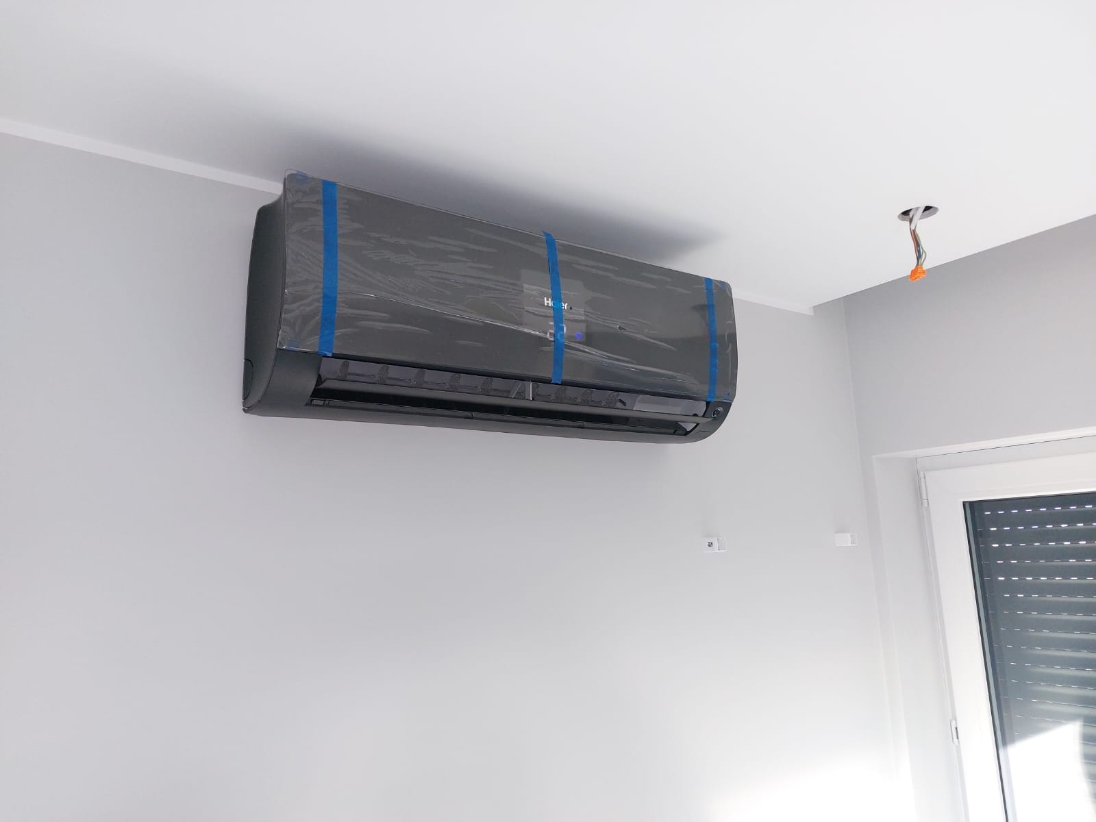 Czarna jednostka klimatyzacji Haier zamontowana na szarej ścianie, zabezpieczona folią i niebieską taśmą, widoczny otwór po instalacji elektrycznej.