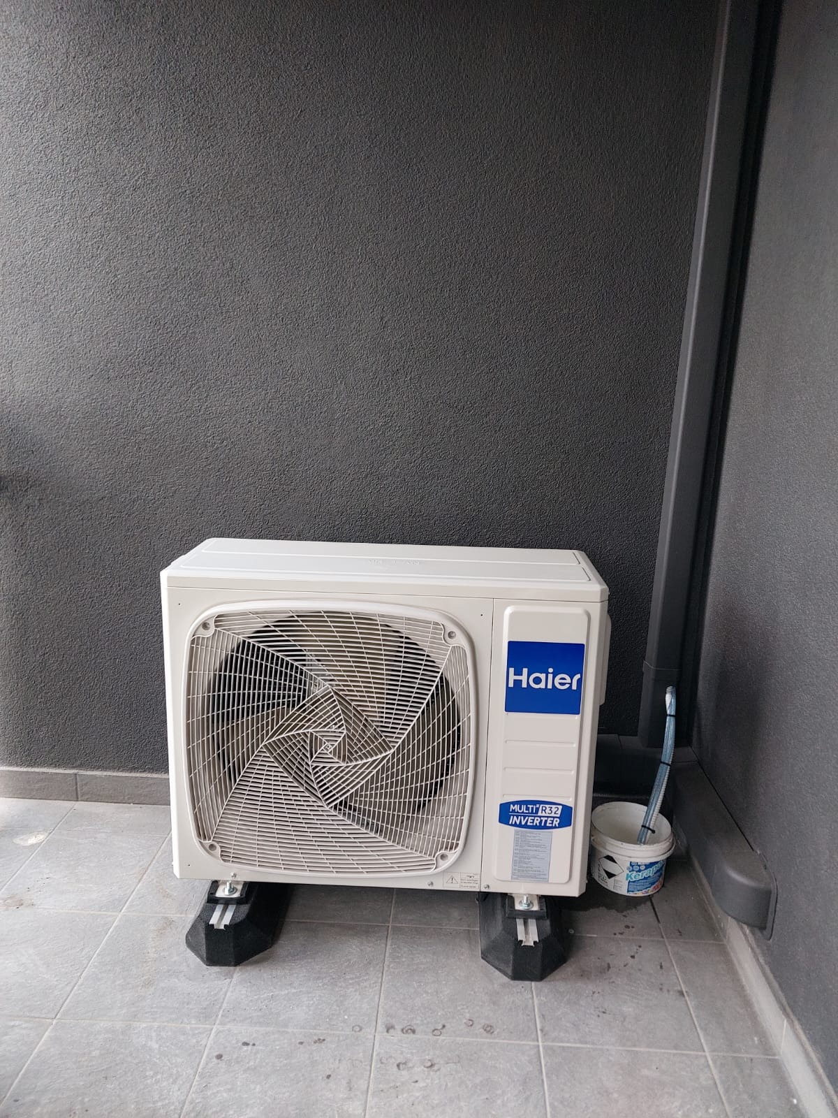 Biała jednostka zewnętrzna klimatyzacji Haier zamontowana na czarnych wspornikach, widoczna puszka z pędzlem malarskim, szara ściana w tle.