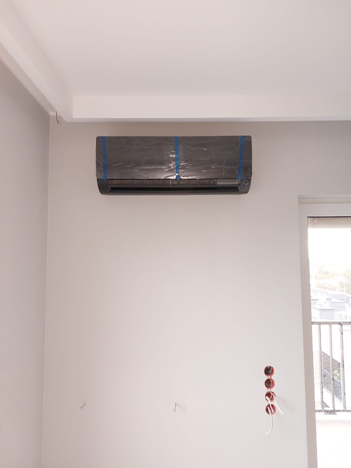 Czarny klimatyzator Haier, jeszcze w folii ochronnej z niebieską taśmą, zamontowany na białej ścianie w jasnym pomieszczeniu, widoczne niedokończone gniazdka elektryczne.