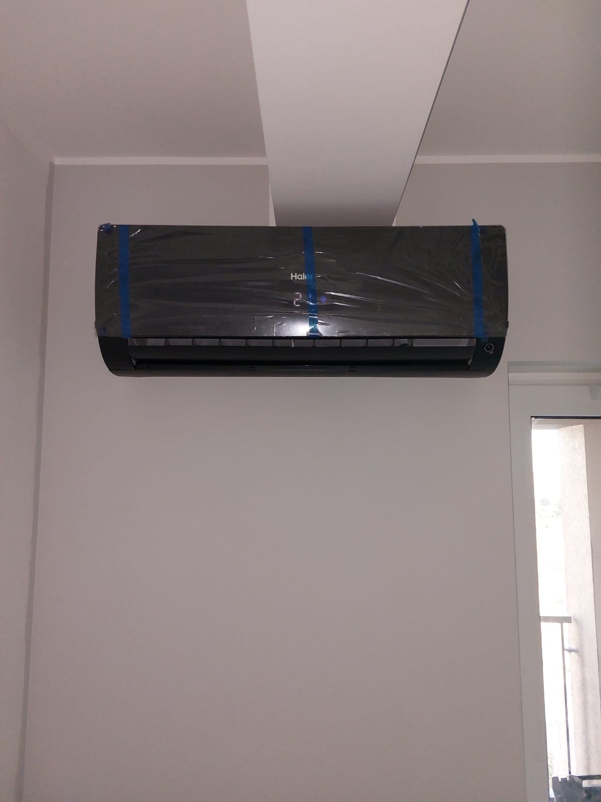 Czarny klimatyzator Haier, jeszcze w folii ochronnej z niebieską taśmą, zamontowany na białej ścianie w pomieszczeniu. Widoczne fragmenty sufitu i okna.