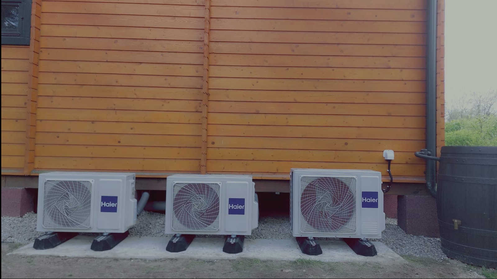 Trzy jednostki zewnętrzne klimatyzacji Haier ustawione rzędem przed drewnianym domem z widoczną beczką na deszczówkę po prawej stronie.