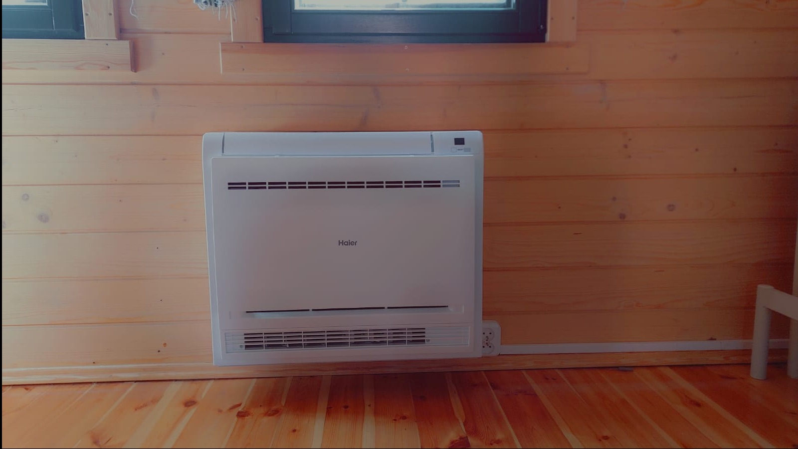 Biała, niskopodłogowa jednostka klimatyzacyjna Haier zamontowana na drewnianej ścianie w domu, widoczne gniazdko elektryczne obok urządzenia.