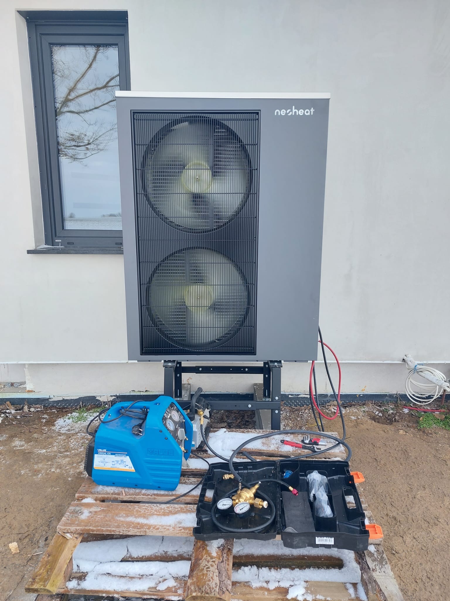 Szara jednostka zewnętrzna z dwoma wentylatorami marki Nesheat zamontowana na metalowym stelażu przy białej ścianie budynku z oknem, z narzędziami do podłączenia na palecie w zimowej scenerii.