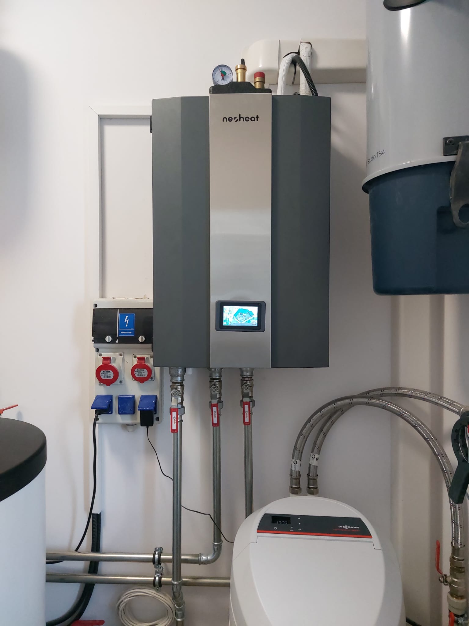 Szary kondensacyjny kocioł gazowy Nesheat z wyświetlaczem, podłączony do instalacji grzewczej Viessmann, widoczne rury doprowadzające wodę i gaz oraz skrzynka elektryczna z gniazdami 400V