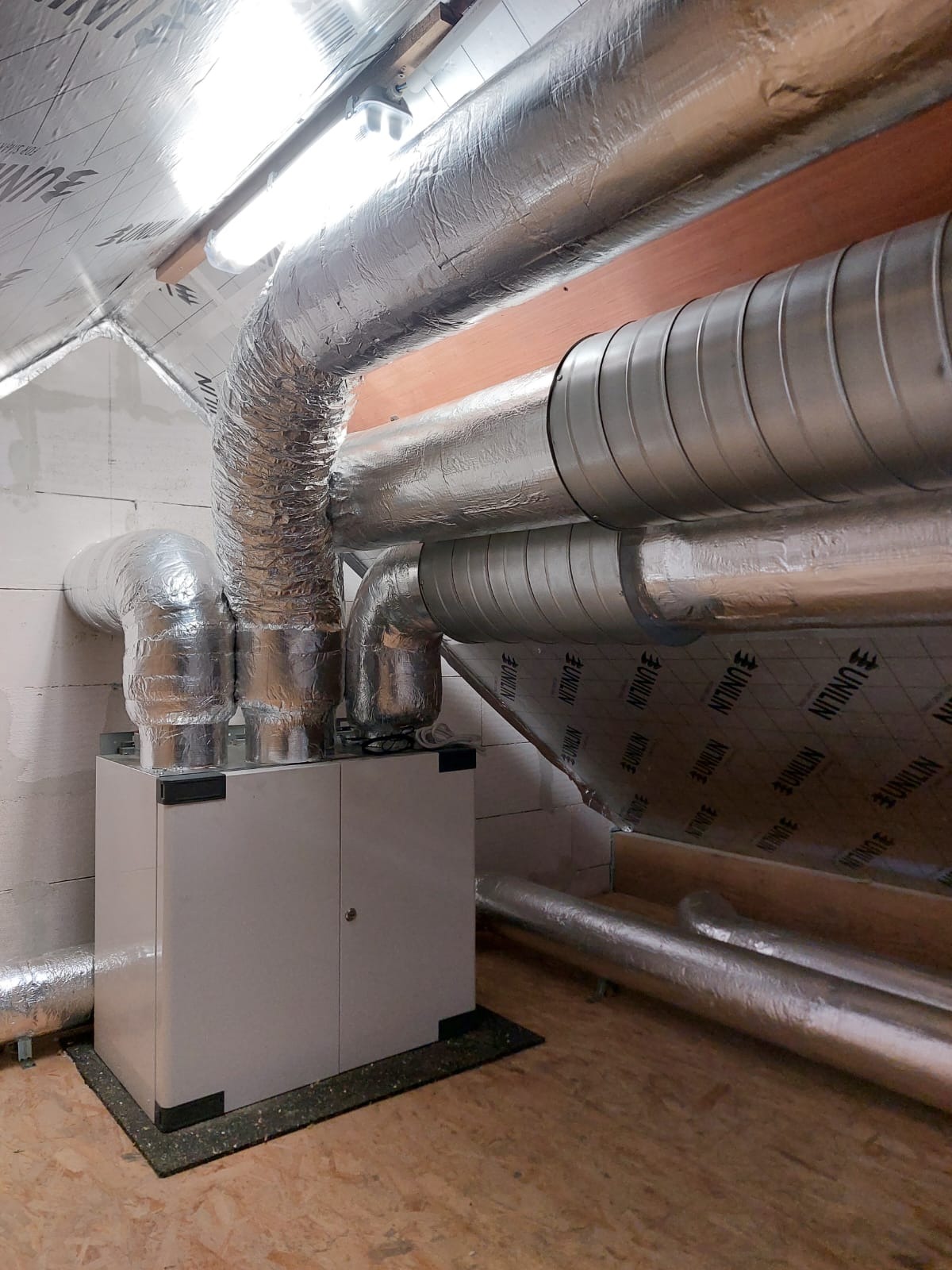 Centrala wentylacyjna z izolowanymi przewodami w pomieszczeniu z wykończeniem z płyty OSB i izolacją, widoczne rury wentylacyjne z folią aluminiową.