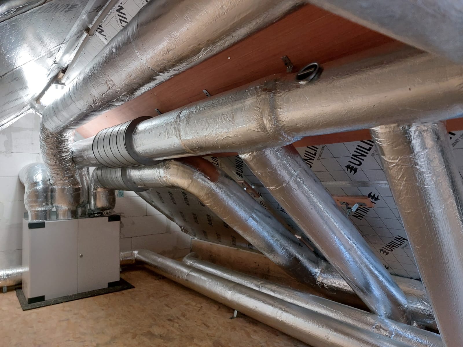 System kanałów wentylacyjnych z centralą rekuperacyjną w nowym budynku, srebrna izolacja na rurach, widoczne elementy montażowe.