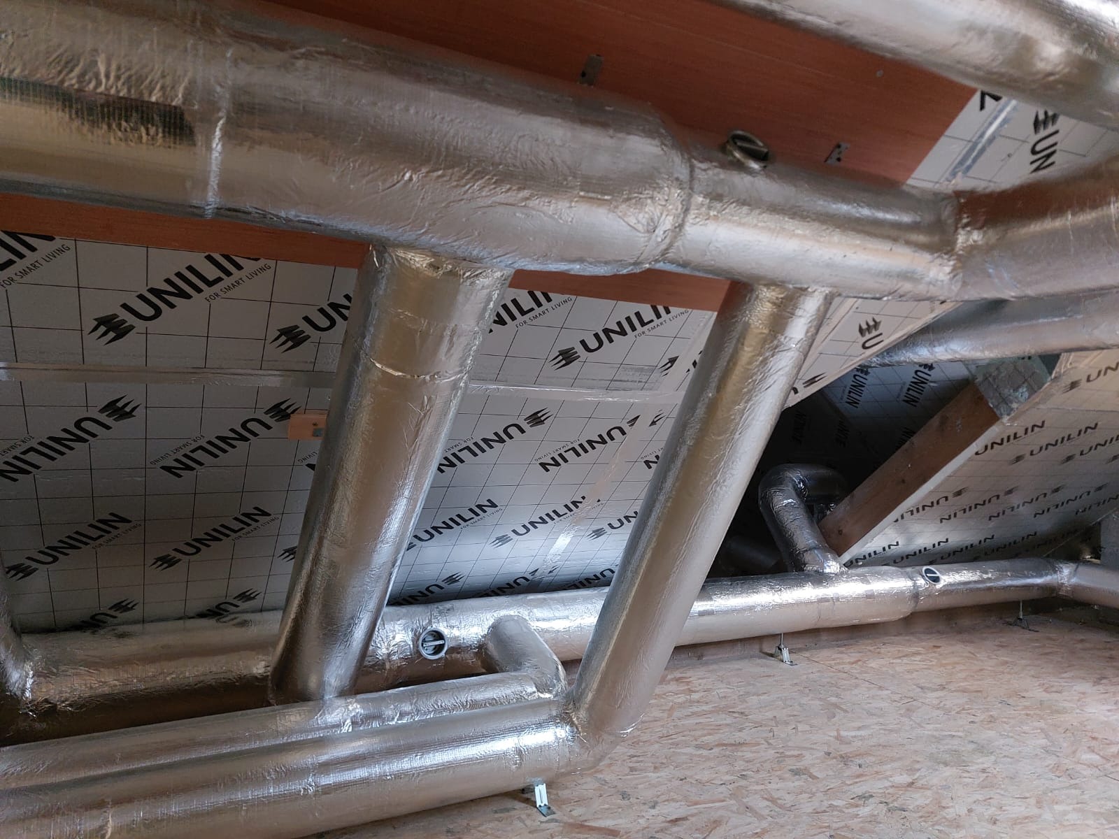 Izolowane rury wentylacyjne w przestrzeni poddasza, widoczne elementy konstrukcyjne dachu i izolacja z nadrukiem logo Unilin.