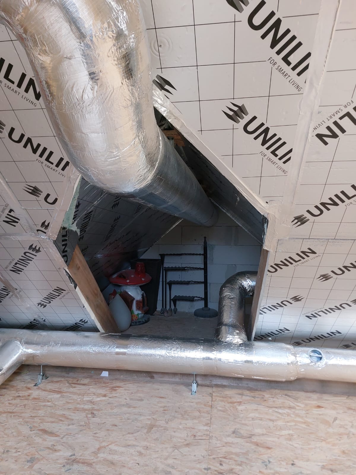 Izolowane rury wentylacyjne w przestrzeni poddasza, widoczne elementy konstrukcyjne dachu i izolacja Unilin, w tle dekoracyjny grzybek i metalowy regał.