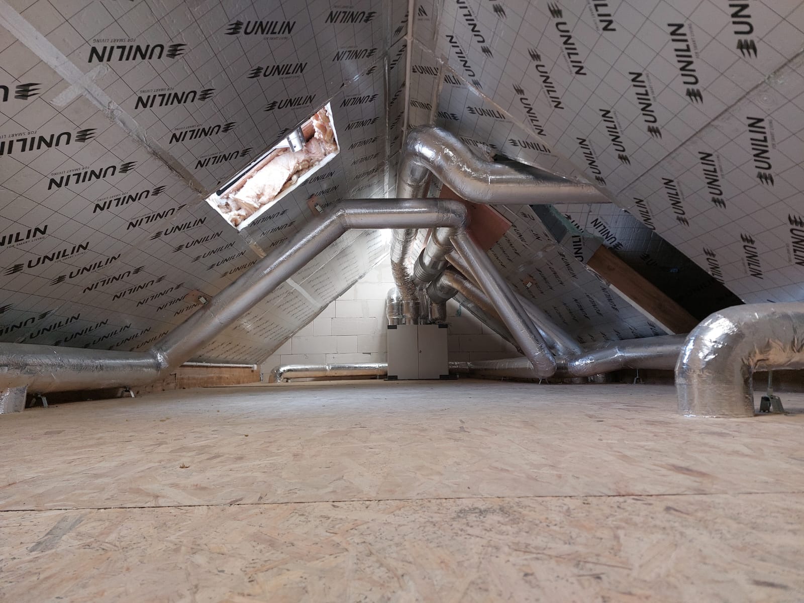 Instalacja rekuperacji na poddaszu z widocznymi rurami wentylacyjnymi, centralą i izolacją Unilin na skośnym dachu.