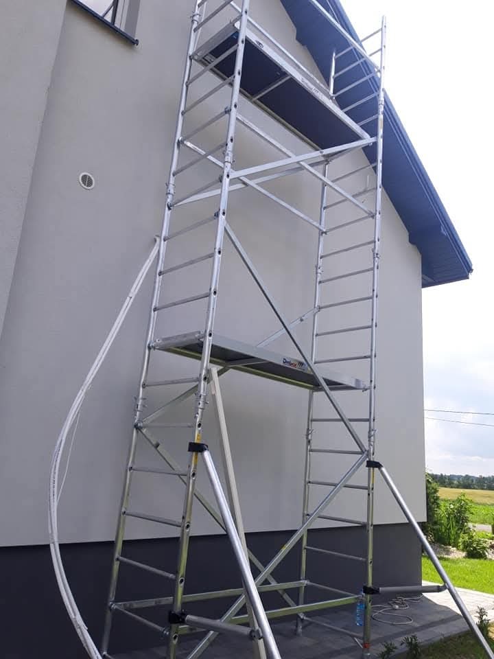 Aluminiowe rusztowanie ustawione przy ścianie domu z widoczną kratką wentylacyjną, prawdopodobnie podczas montażu wentylacji.