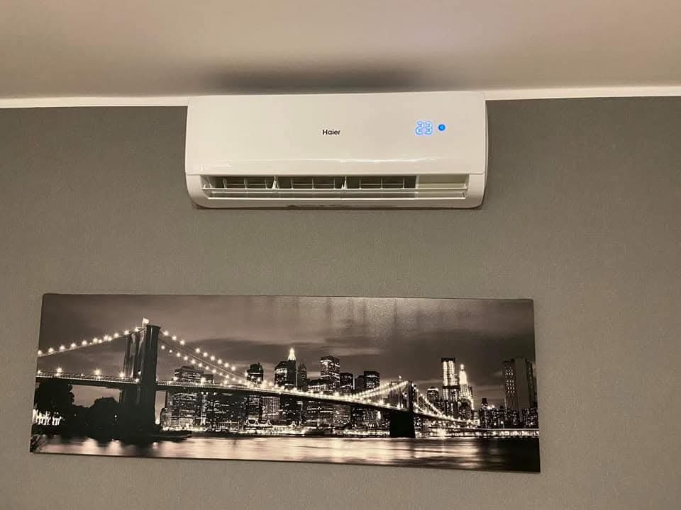 Biały klimatyzator Haier z wyświetlaną temperaturą 23 stopnie, zamontowany na szarej ścianie nad obrazem przedstawiającym panoramę miasta z mostem.