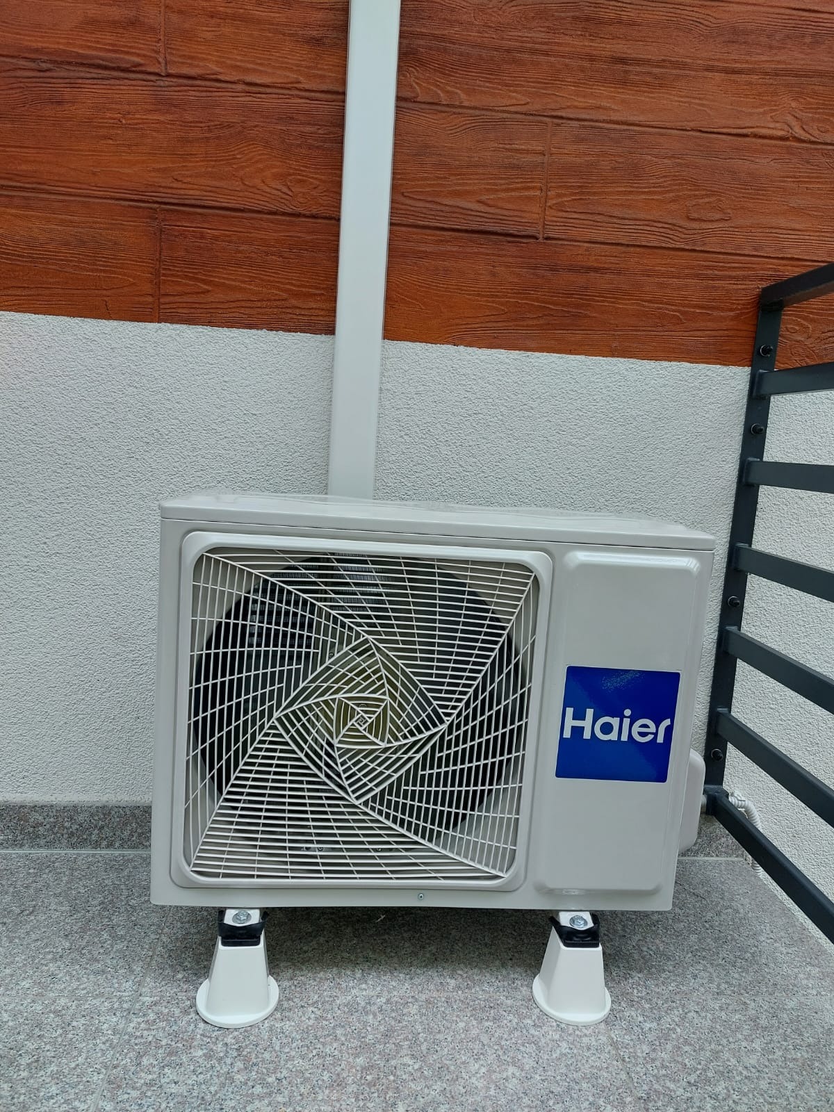 Zewnętrzna jednostka klimatyzacji Haier zamontowana na białych podstawkach, z widoczną instalacją odprowadzającą, na tle ściany z imitacją drewnianej boazerii.