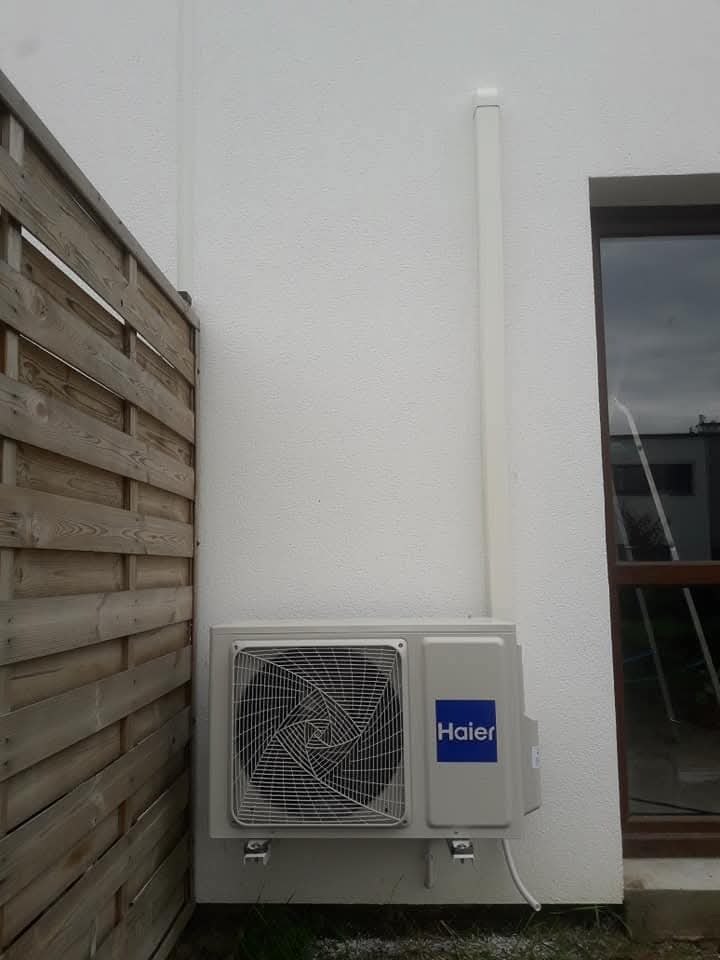 Zamontowana jednostka zewnętrzna klimatyzatora Haier przy białej ścianie budynku, widoczna listwa maskująca instalację, obok drewniany płot i okno.