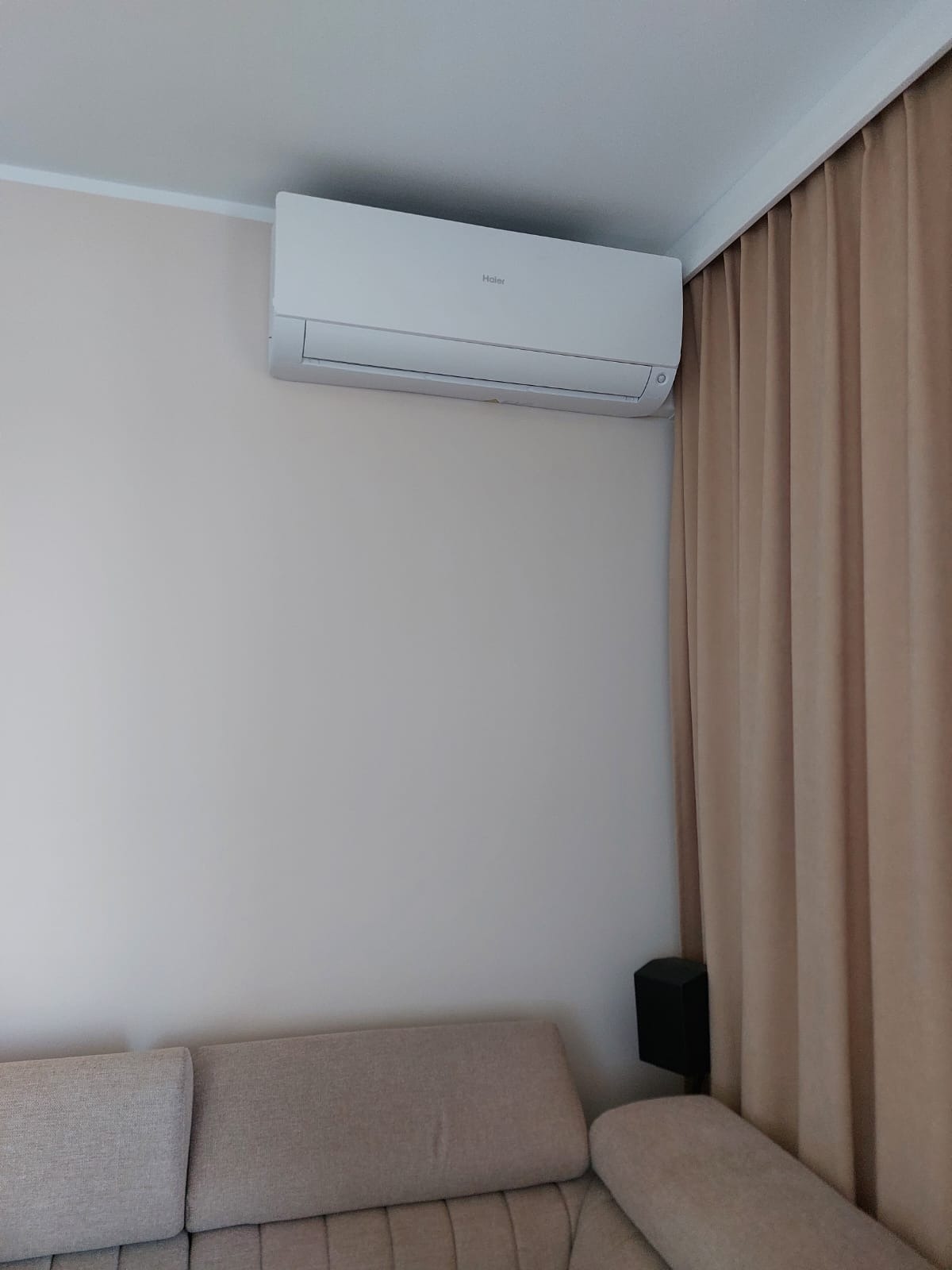 Biała jednostka klimatyzacji Haier zamontowana na ścianie nad beżową sofą, obok zasłon w kolorze piasku.