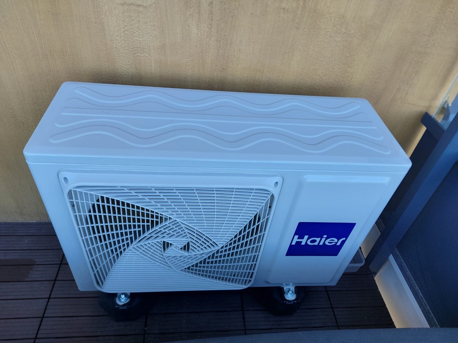 Biała jednostka zewnętrzna klimatyzacji Haier ustawiona na kółkach, widoczna z boku na tle beżowej ściany i fragmentu czarnej barierki.