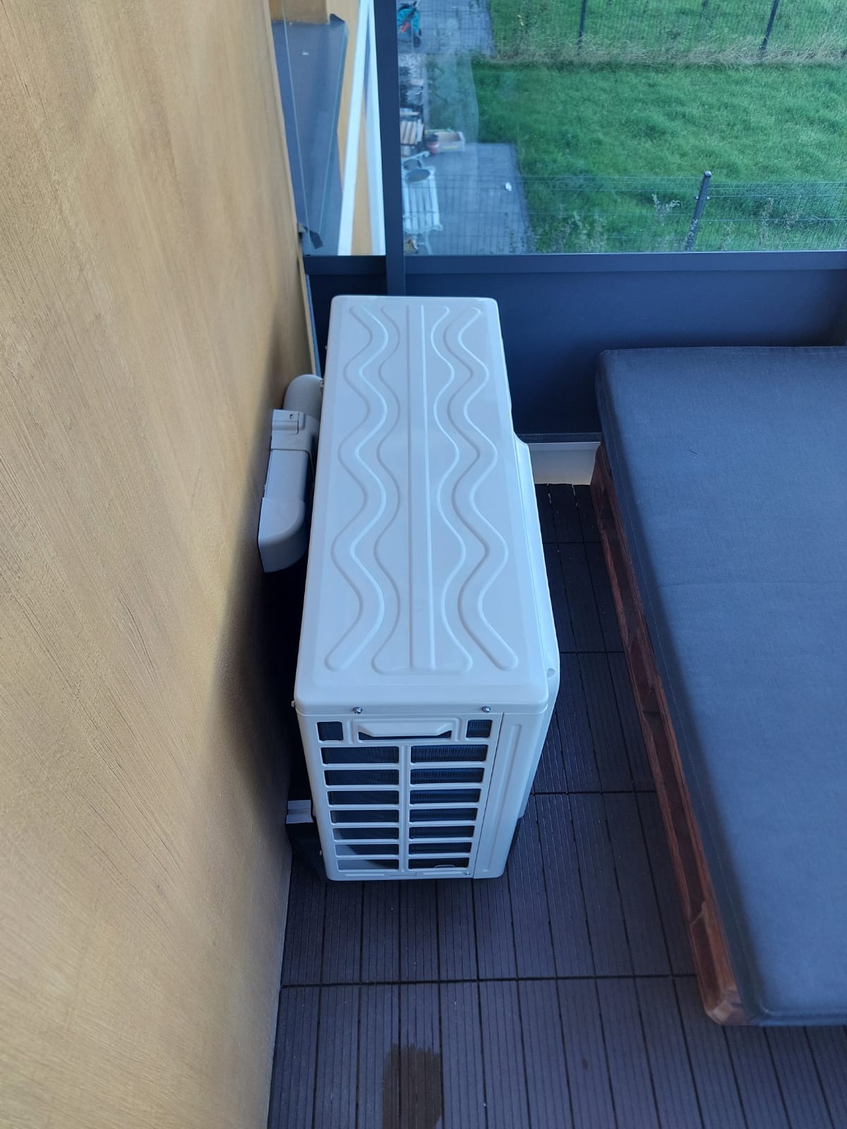 Biała jednostka zewnętrzna klimatyzatora zamontowana na balkonie, widoczne przewody odprowadzające skropliny, obok leżanka z szarą poduszką.