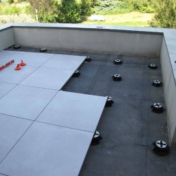 WAWER-BRUK Mariusz Wiącek - Montaż tarasu wentylowanego: jasnoszare płytki układane na regulowanych wspornikach, poziomica i przyssawki do płytek obok, widok z góry na częściowo wykończony taras.